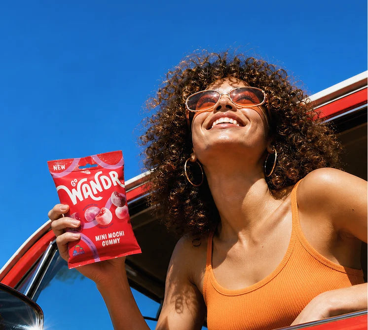 A smiling woman with curly hair and sunglasses leans out of a red car under a bright blue sky, holding Go Wandr mini mochi gummies