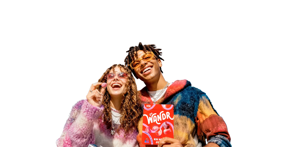 Two people smiling and posing together against a solid black background, wearing colorful fuzzy sweaters and sunglasses, with one holding Go Wandr mini mochi gummies