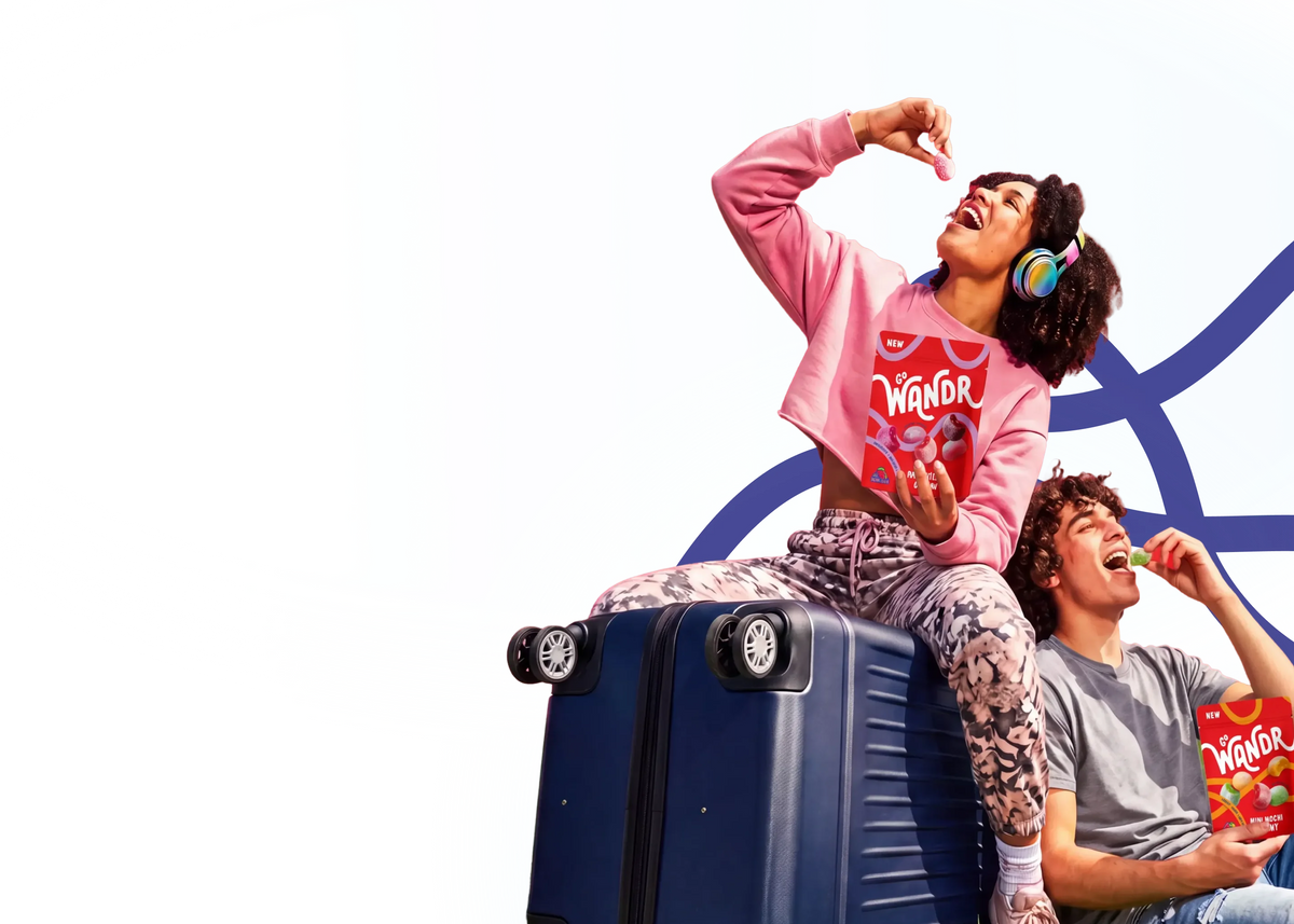 A woman in a pink sweatshirt and patterned pants sits on top of a blue suitcase, eating snacks while wearing headphones. A man in a gray shirt is seated beside her, also eating and holding Go Wandr mini mochi gummies